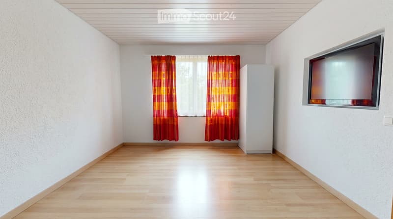 Empty room with white walls, wooden floor, and a mounted TV on one wall. Red and yellow curtains cover a window, and a small white cabinet is positioned near the window.