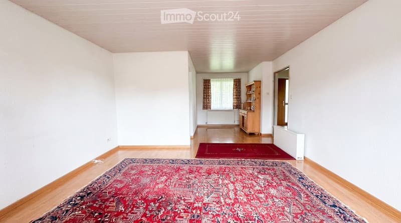 Bright hallway with wooden flooring, white walls, and two area rugs leading to a kitchen area with wooden cabinets and a window.