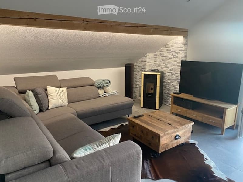 Modern living room with a grey sectional sofa, wooden coffee table, flat-screen TV on a wooden stand, and a built-in fireplace. The room features exposed wooden beams and a textured wall finish.