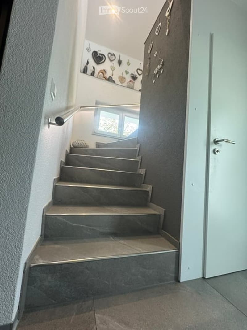 Modern staircase with grey stone steps, metal railing, and a small window at the top. The walls are painted in neutral tones with decorative wall art and a small shelf. The lighting is bright, and the overall design is contemporary.