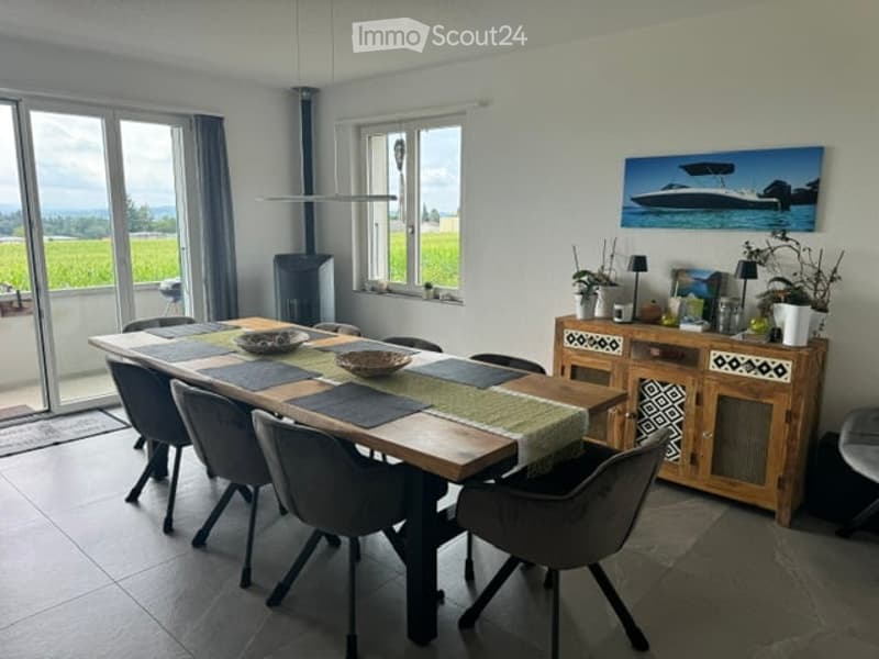 Bright dining room with large windows offering a scenic view, a wooden dining table surrounded by modern chairs, and a wooden cabinet with decorative items.