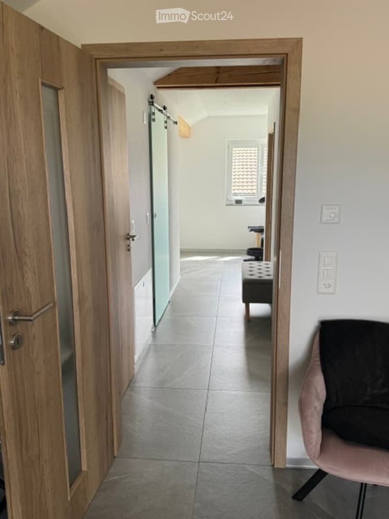A modern hallway with wooden doors, tiled floor, and a glimpse of a room with a pink chair and a small table in the background.