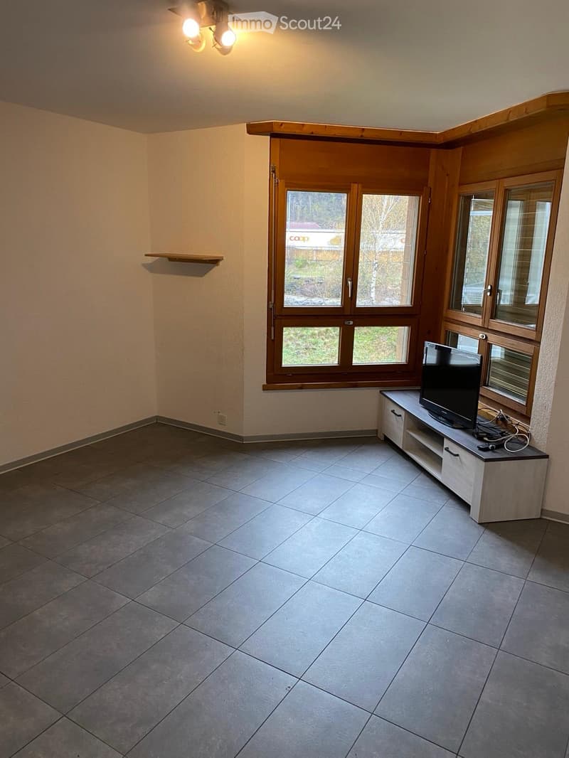 Empty room with tiled floor, large windows, TV stand with television, and a small shelf on the wall.