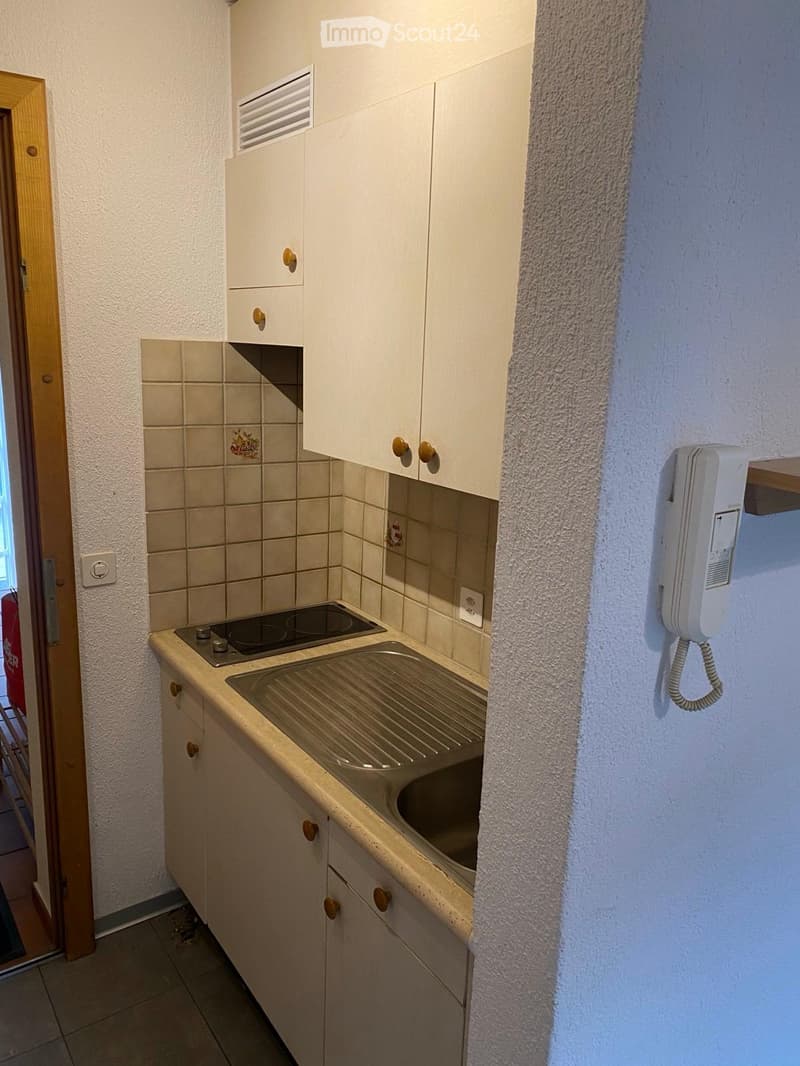 Compact kitchen with white cabinets, beige countertop, sink, and electric stove. Features tiled backsplash and a telephone mounted on the wall.