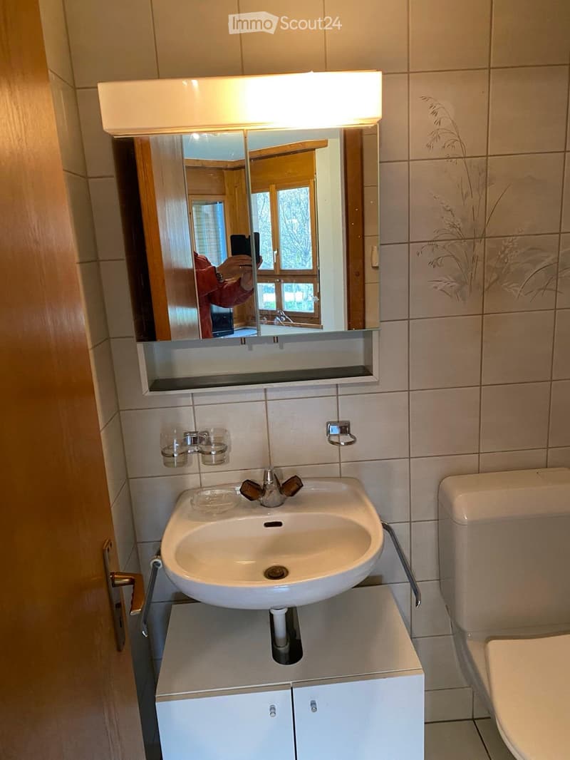 Modern bathroom with a white sink, mirror with built-in lighting, and a toilet. The walls are tiled, and there is a small cabinet under the sink.