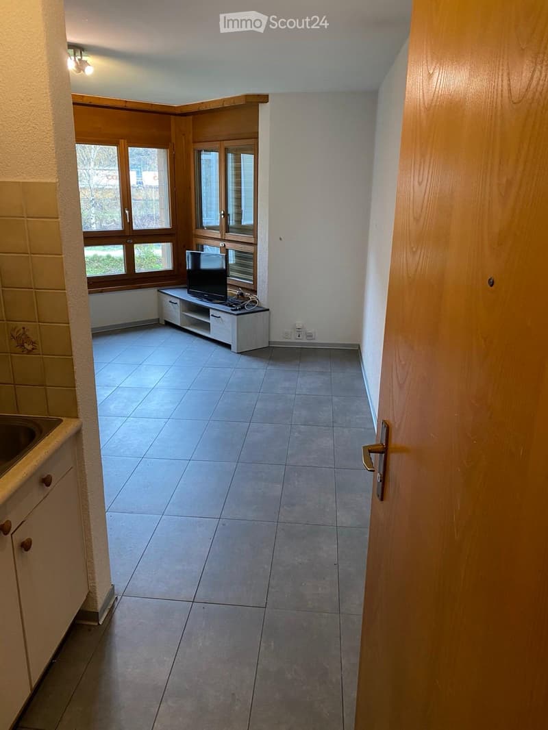 A small, unfurnished room with tiled flooring, a TV stand with a screen, and a sink visible in the background.