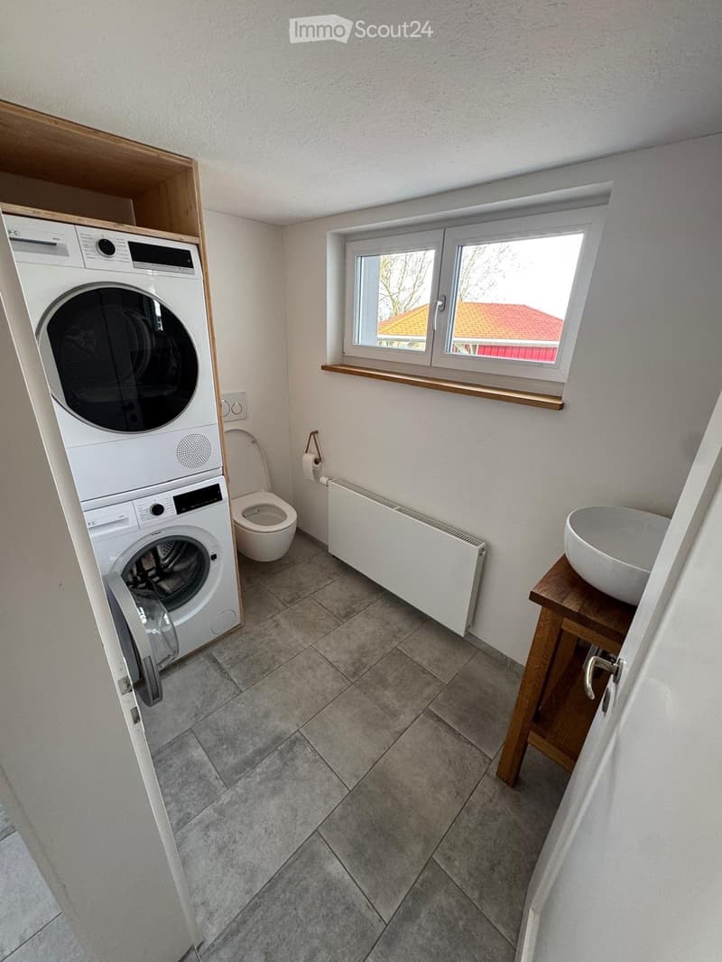 Bathroom with tiled floor, white walls, a washing machine, toilet, sink, and a window with a wooden sill.
