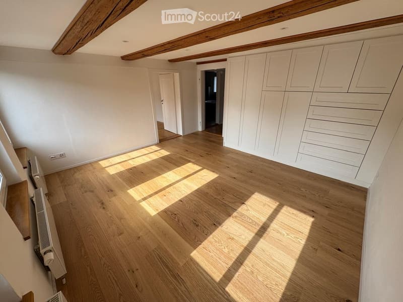Empty room with light wooden flooring, white walls, built-in white cabinets, and exposed wooden beams on the ceiling. Sunlight streams in from a window, casting shadows on the floor.