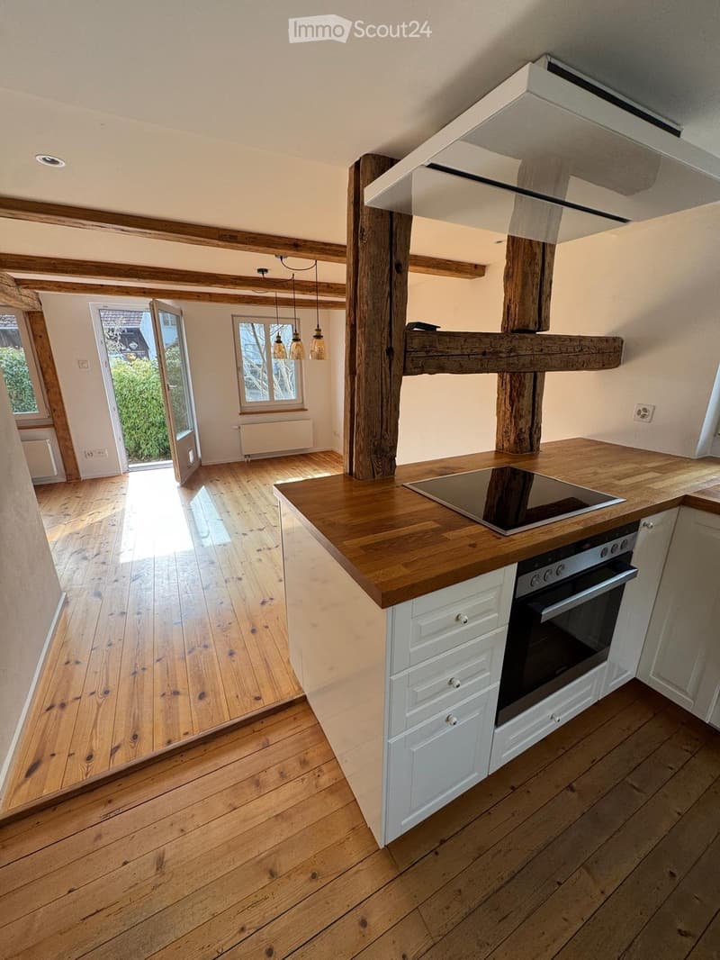 Modern kitchen with wooden countertops, white cabinets, induction stove, and pendant lights. Features exposed wooden beams and a large window with a view of greenery.
