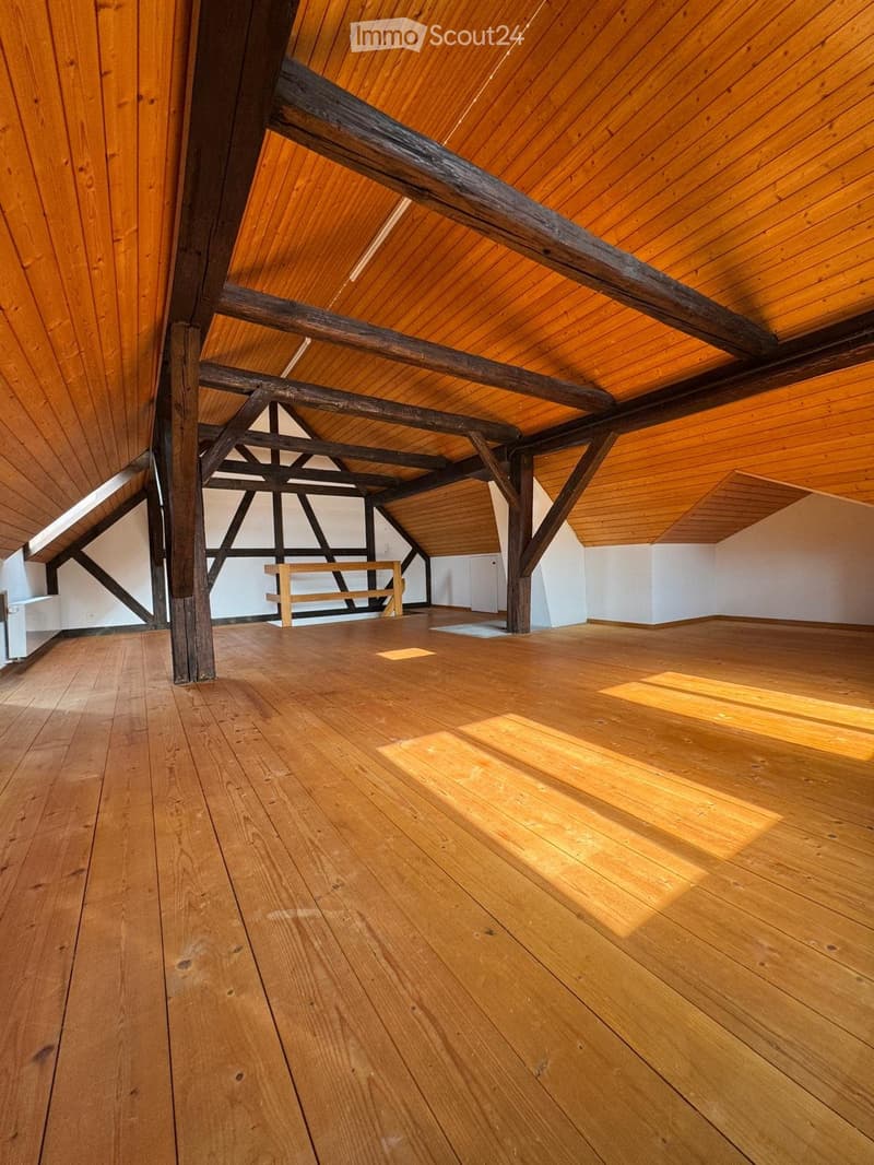 Empty attic space with wooden floors and exposed wooden beams, white walls, and a small wooden bench in the background.