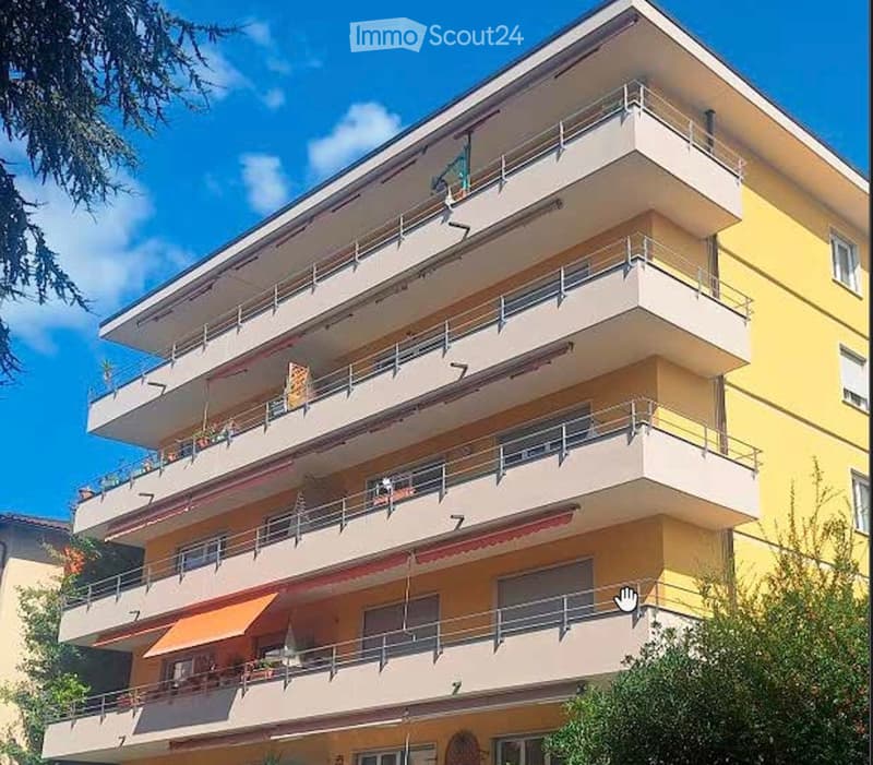 Multi-story residential building with balconies, yellow and beige facade, and visible potted plants on the balconies.