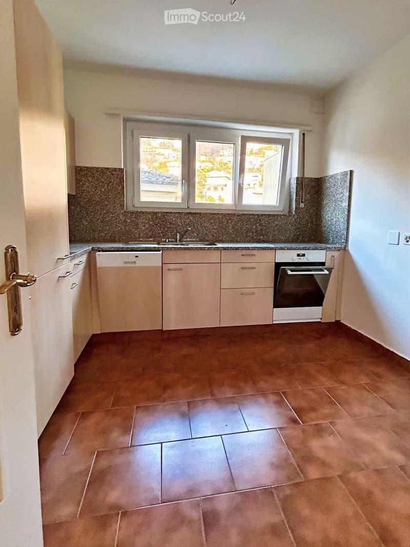 Empty kitchen with light-colored cabinets, a sink, a dishwasher, and an oven. The floor is tiled in a warm brown color, and the backsplash is made of a speckled gray material.
