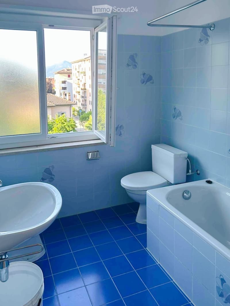 Bathroom with blue tiles, white fixtures, and a view of the city through a window.