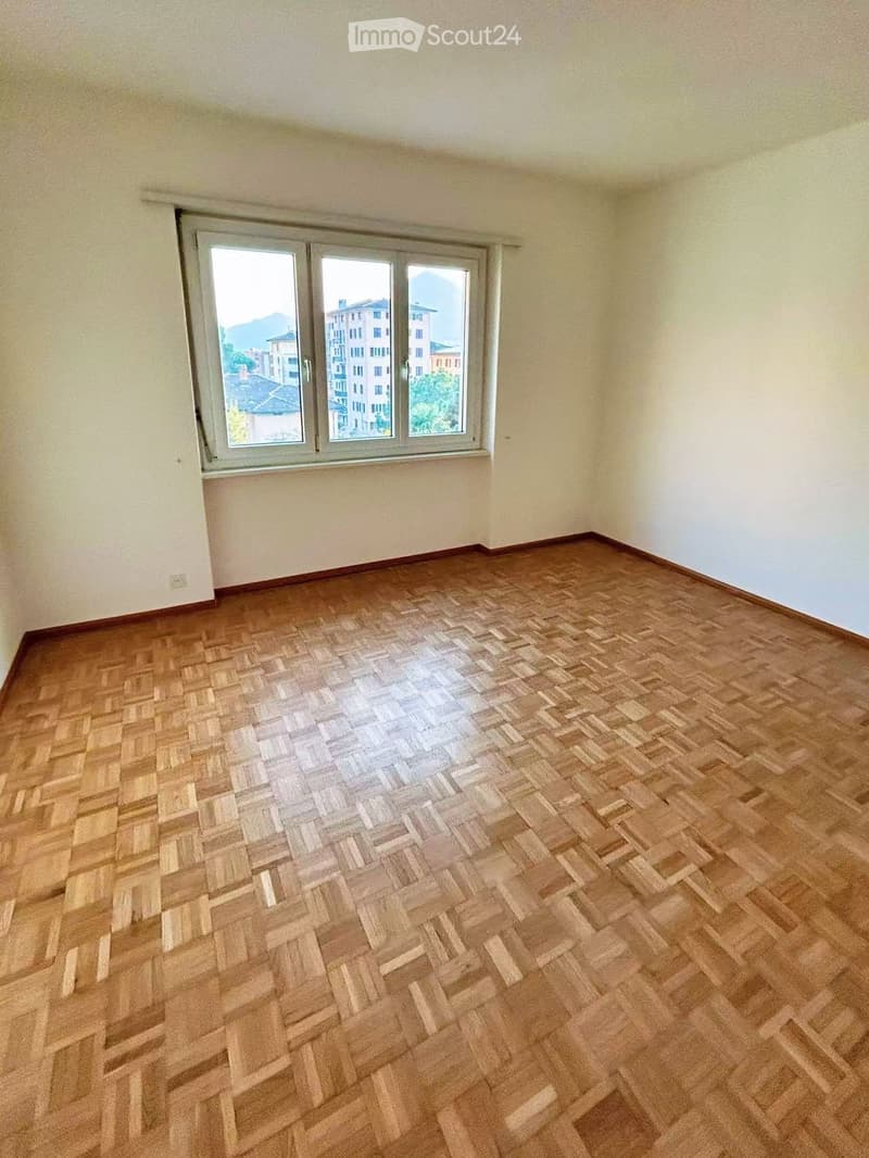 Empty room with wooden herringbone flooring and large windows offering a view of buildings and mountains.