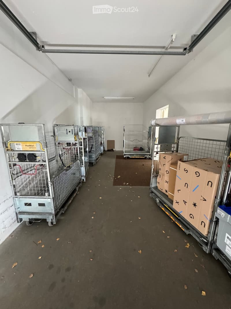 Empty garage with metal cages and storage boxes, concrete floor, white walls, and a small window at the far end.