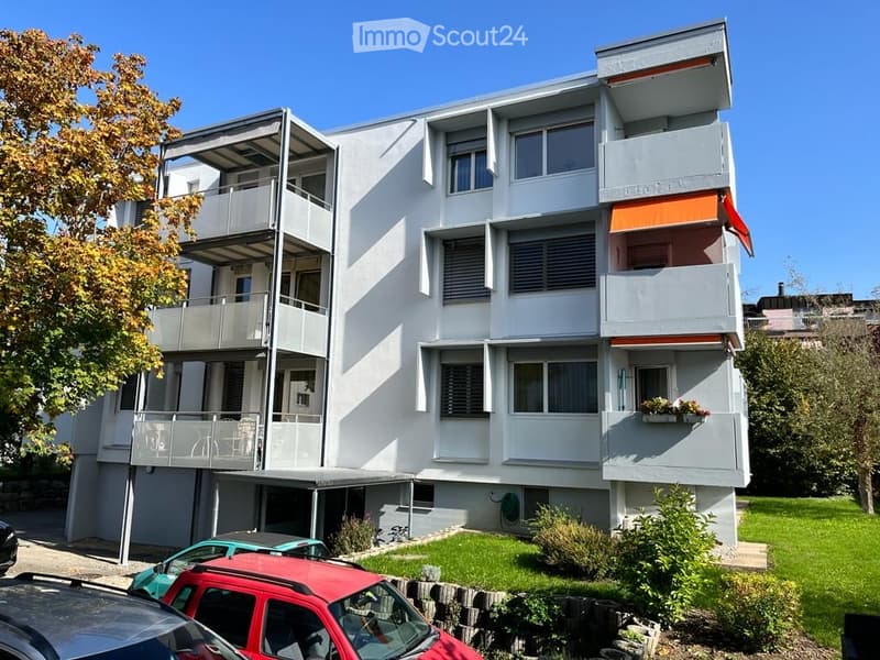 Modern multi-story apartment building with white facade, multiple balconies, and a small garden area with parked cars.