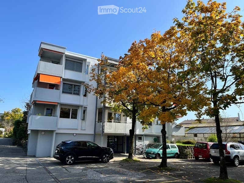 Modern multi-story residential building with balconies, surrounded by trees and parked cars.