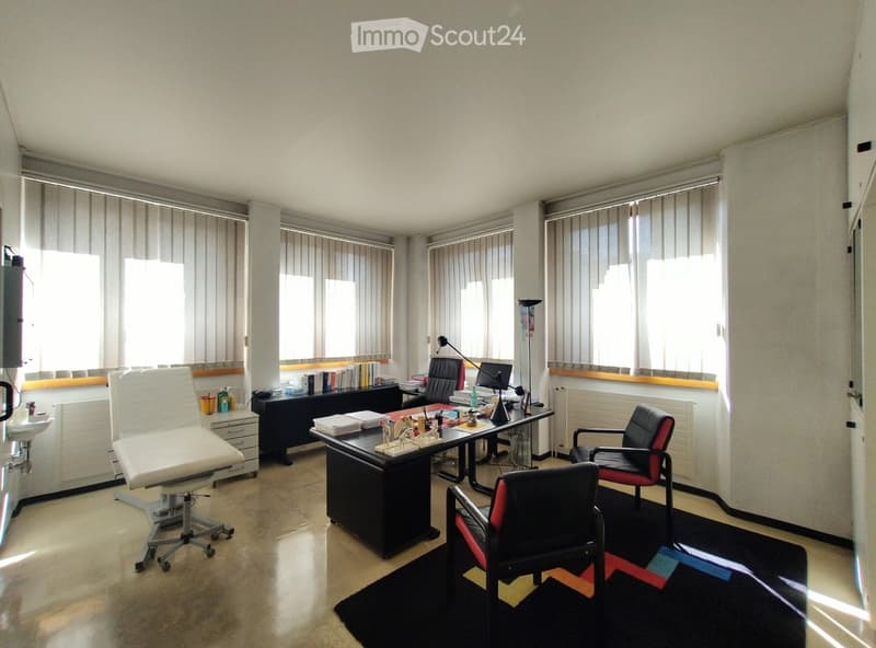 Office space with a large desk, chairs, a computer screen, a lamp, and a bookshelf with books and office supplies.