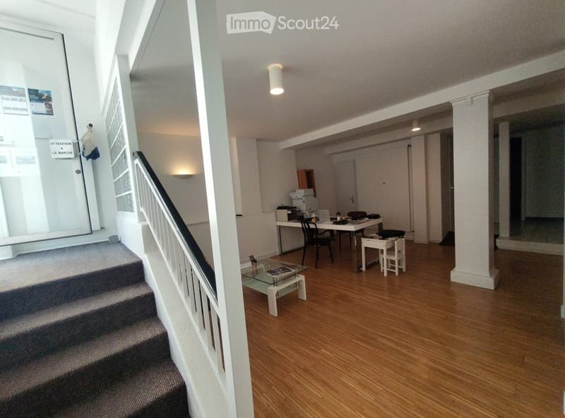 Spacious, well-lit room with wooden flooring, a staircase leading upwards, a coffee table, a desk with a laptop, and a small seating area with a chair and stool.