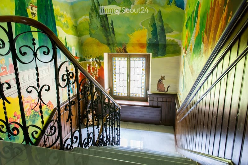 Staircase with ornate wrought-iron railing, colorful mural on walls, stained-glass window at landing, small wooden bench, and a cat figurine on a cabinet.