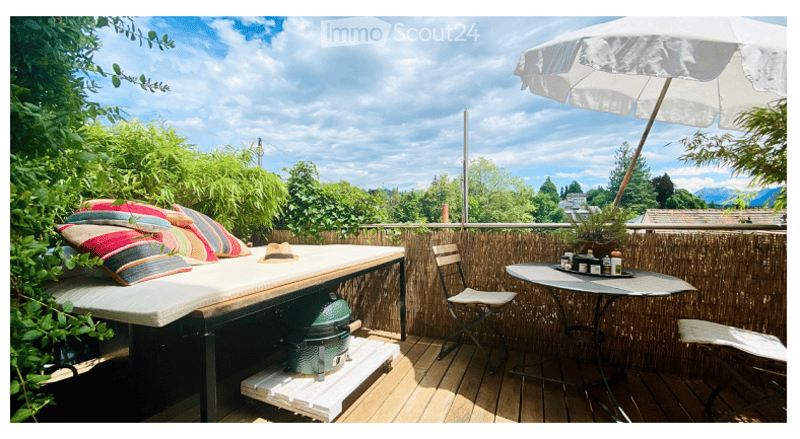 Balcony with wooden decking, outdoor furniture including a daybed with colorful pillows, a small table with chairs, and a large umbrella providing shade. The background shows a scenic view of trees and mountains.