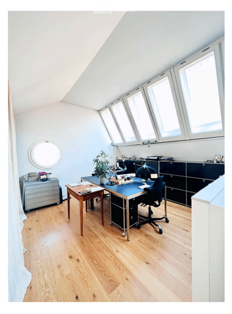 Bright, modern office space with wooden flooring, large windows, a desk, chair, cabinet, and a small plant. The room features a slanted ceiling and ample natural light.