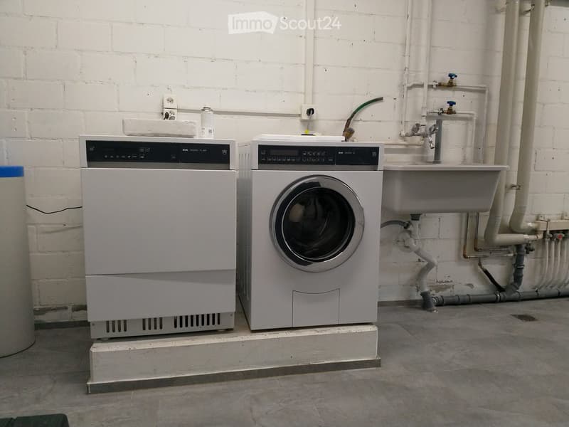 A laundry room with white walls and floor, featuring a washing machine and dryer next to a sink.