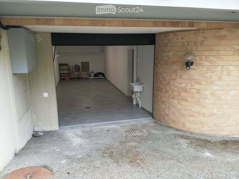 Empty garage with tiled floor and a small sink. Open garage door and a lamp mounted on the wall