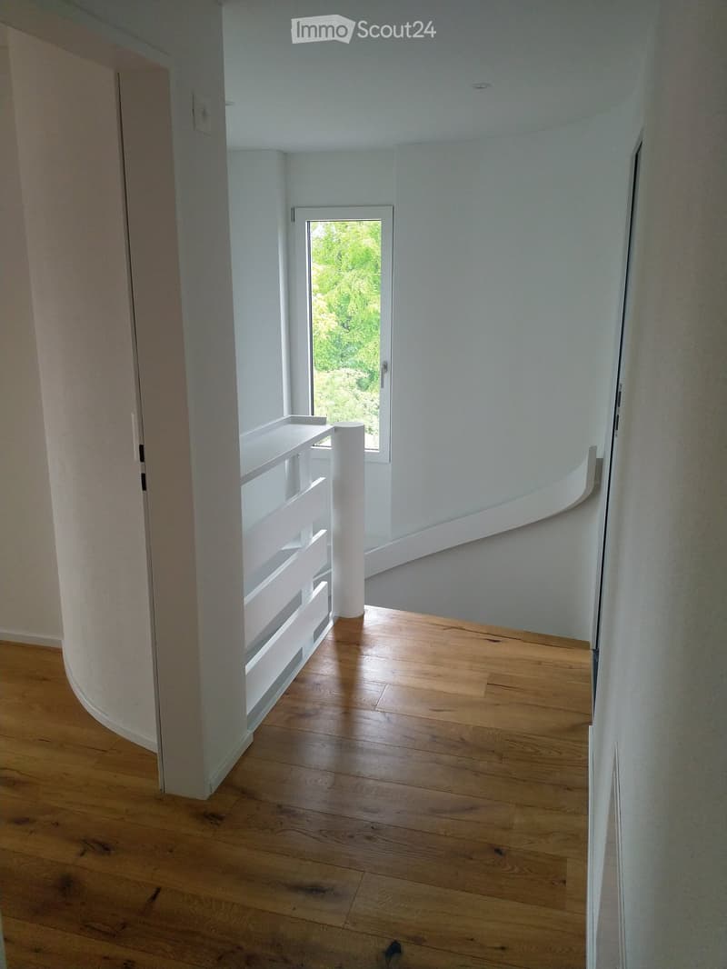 wide entrance hall with wooden flooring, curved staircase with white railing, white wall, and a window.