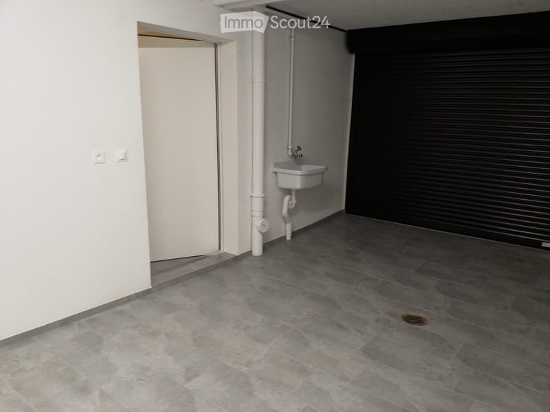 Clean and empty room with gray tiles, white walls, door, sink with faucet, black roll-up door, gray grout, floor drain
