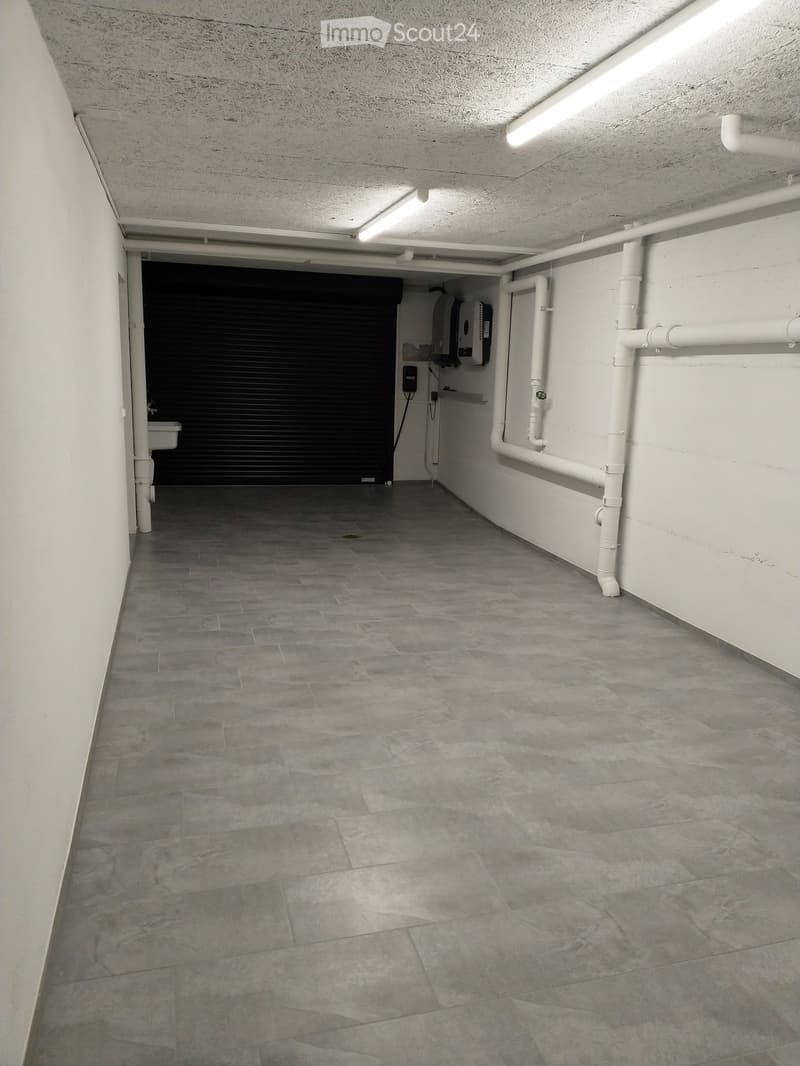 Empty room with tiled flooring, white walls, black roller door, pipe system on wall, sink and light
