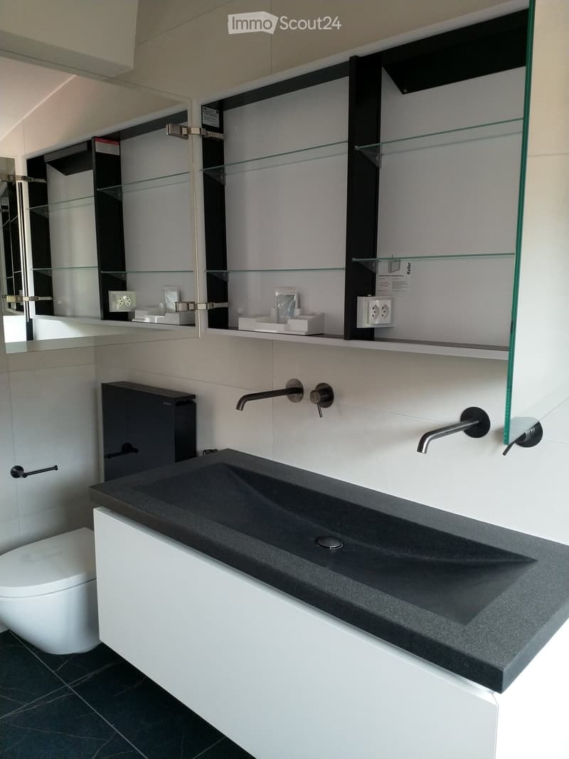 bathroom with white tile walls, black countertops, modern sink, large glass cabinet, toilet, and two black faucets.