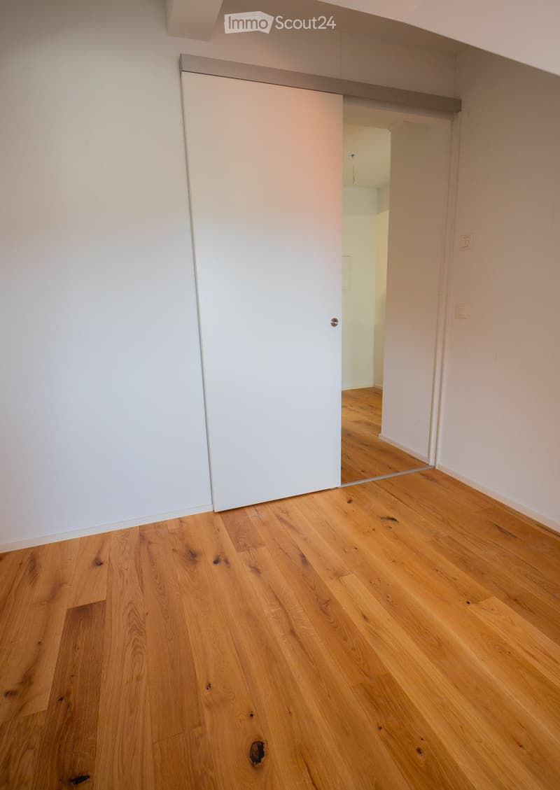 Empty room with light wooden flooring and white walls, featuring a closed white sliding door leading to another room.