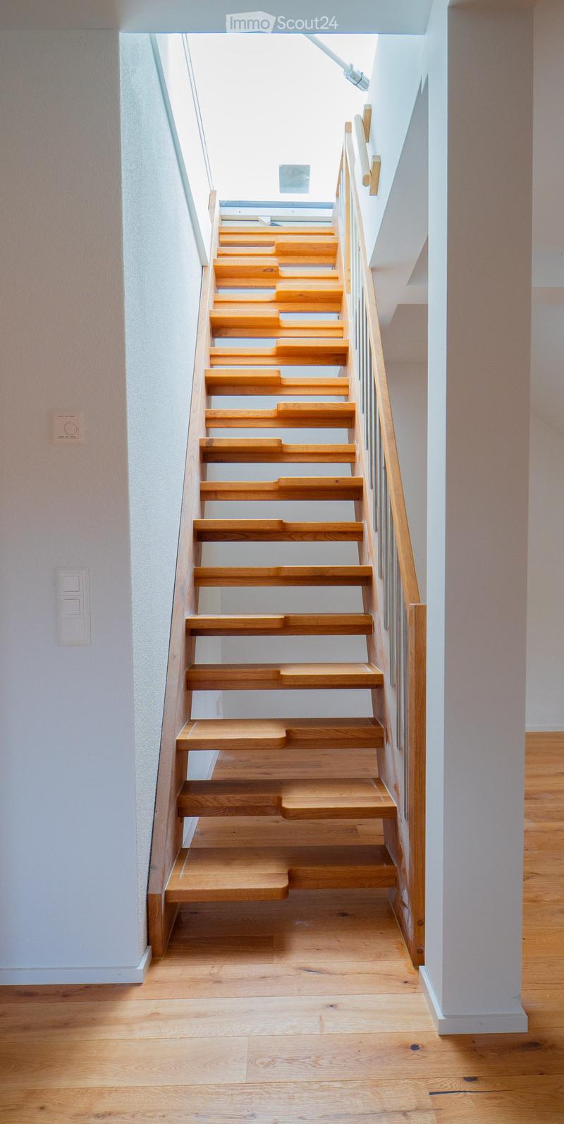 Wooden staircase with light-colored steps and a wooden handrail, set against white walls and a wooden floor.