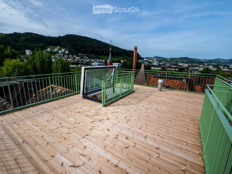 Wooden deck with green metal railings, offering a scenic view of a town and surrounding hills.