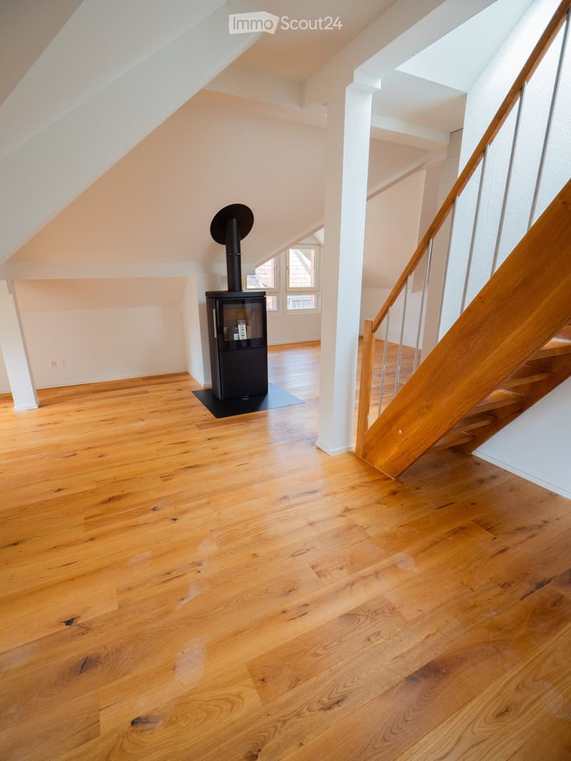 Bright hallway with wooden flooring, white walls, and a modern black wood-burning stove. A wooden staircase with a metal railing leads upwards.