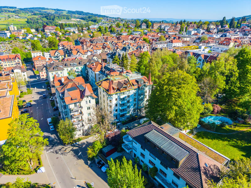 Aerial view of a residential area with multiple buildings, red-tiled roofs, green spaces, and a swimming pool in the foreground.