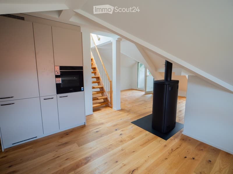 Modern room with light wooden flooring, white cabinetry, built-in oven, staircase leading upwards, and a central black wood-burning stove.