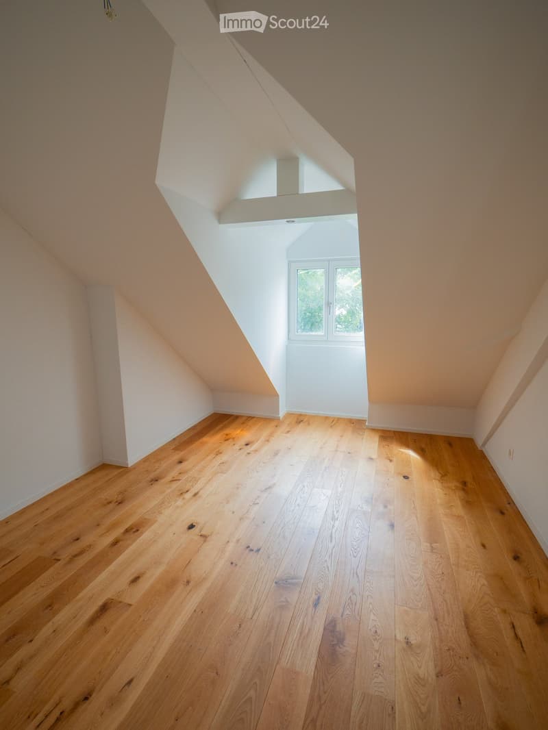 Empty attic space with wooden flooring, slanted ceilings, and a small window letting in natural light.