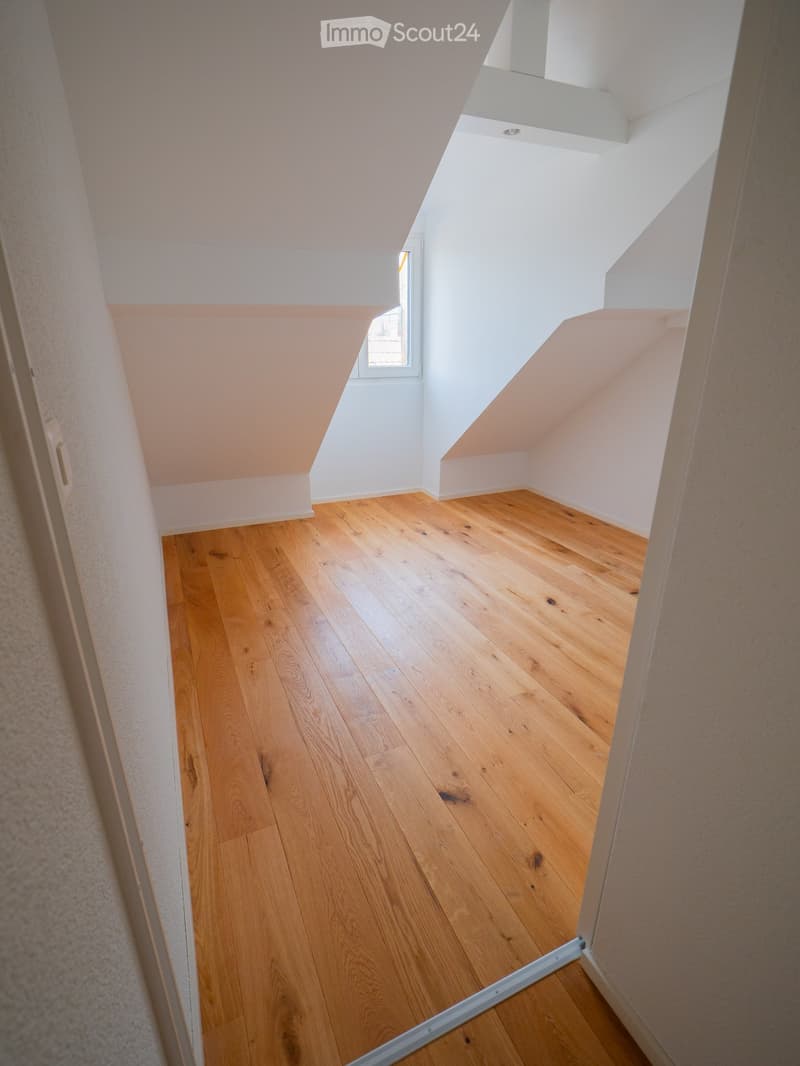 Empty attic space with wooden flooring, white walls, and a small window allowing natural light.
