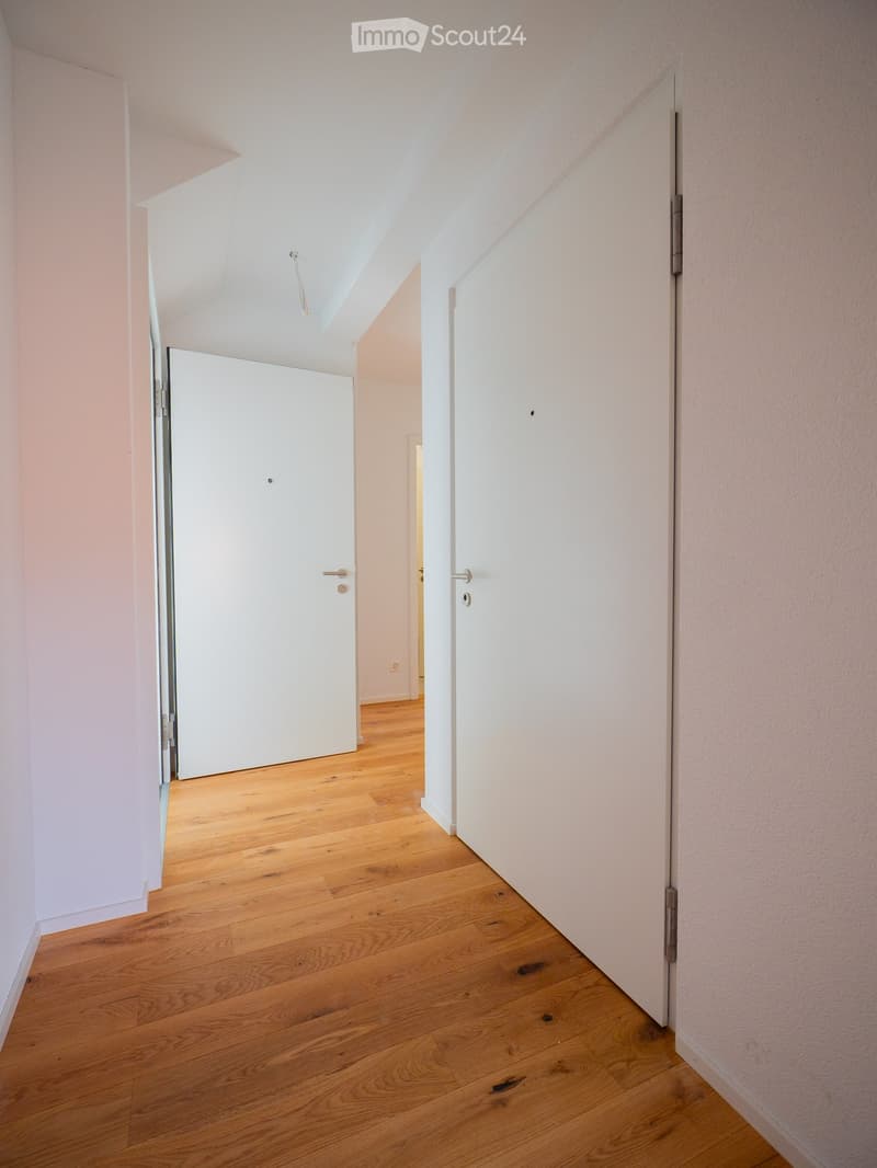 Empty hallway with light wooden flooring and white walls, featuring two closed doors and a visible electrical outlet.