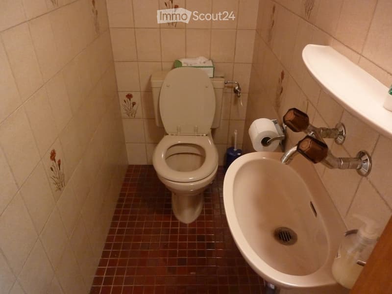 A small bathroom with beige tiled walls and brown tiled flooring. It features a toilet, a sink with a faucet, a toilet paper holder, and a toilet brush holder.