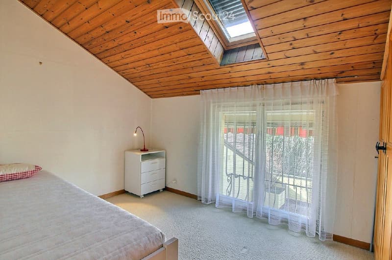 Cozy bedroom with a wooden slanted ceiling, skylight window, bed with a pillow, nightstand with a lamp, and sheer curtains covering a window with a view of a staircase.