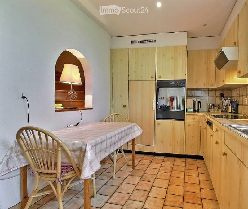A cozy kitchen with light wooden cabinets, a tiled floor, and a dining area featuring a table and chairs. A lamp is placed on a small shelf in an arched alcove.
