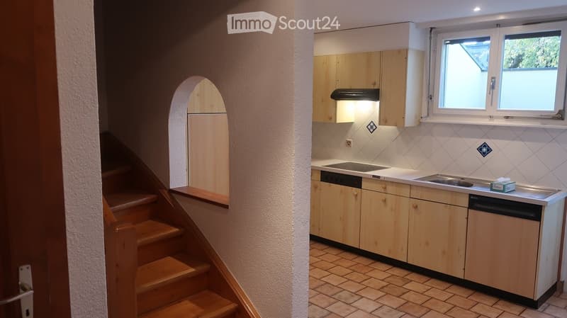 Compact kitchen with light wooden cabinets, a sink, stove, dishwasher, and refrigerator. The space features tiled flooring and a small window above the sink.