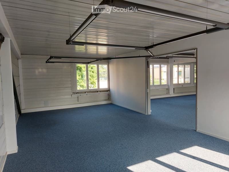 Empty room with blue carpet, white walls, large windows, and a metal ceiling with exposed beams.