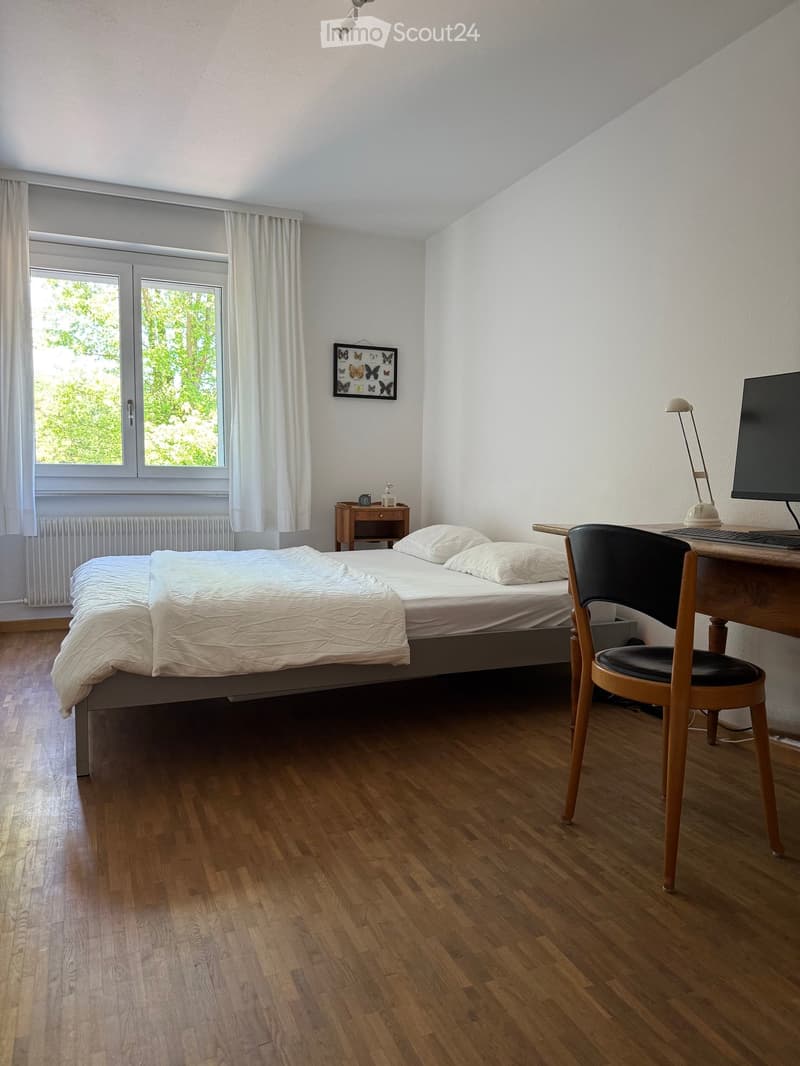 Cozy bedroom with wooden flooring, a neatly made bed, a nightstand, a desk with a computer, and a chair. Large windows with white curtains allow natural light to fill the space.