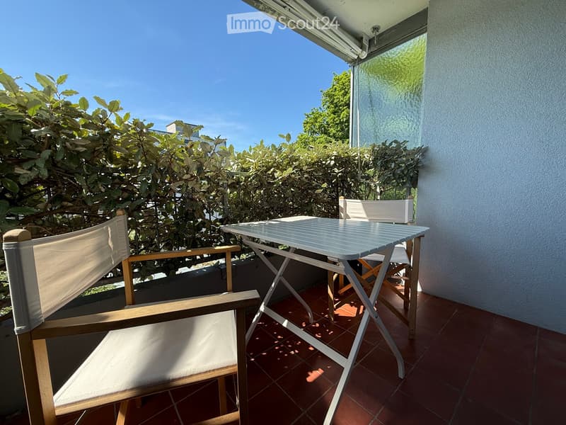 Balcony with a table and two chairs, surrounded by greenery and a clear blue sky.