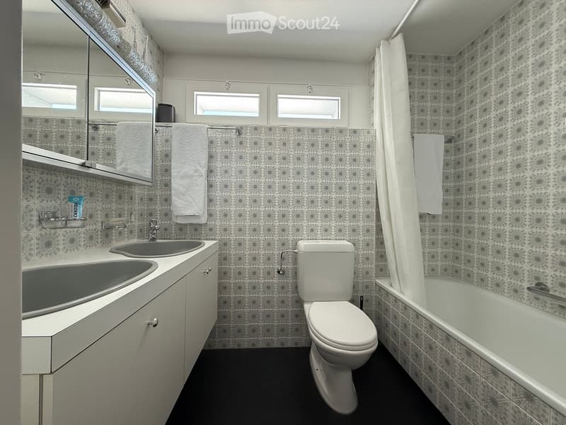 Modern bathroom with double sink, toilet, bathtub, and mirror. Features patterned tiles and white towels.