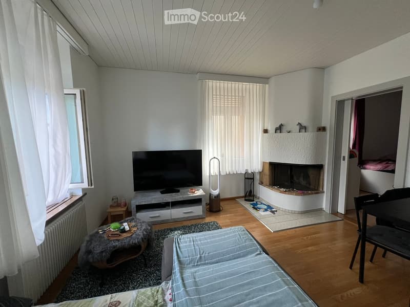 A modern living room with a fireplace, a sofa, a coffee table, a television, and a chair. The room features wooden flooring and large windows with white curtains.
